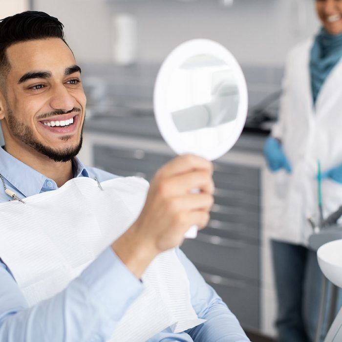 Happy Handsome Arab Man Looking To Mirror After Teeth Treatment In Modern Clinic, Middle Eastern Male Patient Sitting In Chair In Stomatological Cabinet And Enjoying His New Smile, Closeup